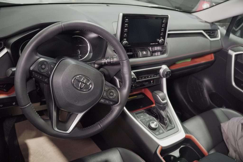 Close-up of a Toyota vehicle interior showing the steering wheel, dashboard touchscreen, gear selector, and control buttons.