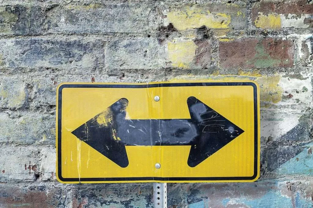 Weathered yellow road sign with black arrows pointing left and right mounted on a brick wall.