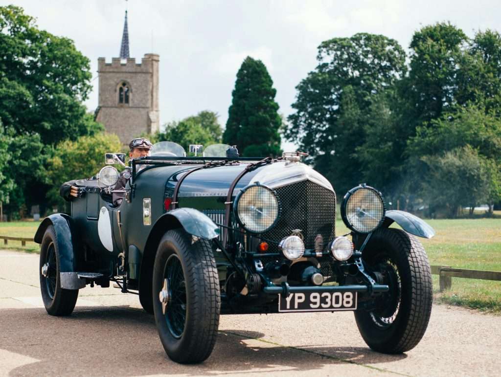 An early motor vehicles driven by a man wearing goggles, featuring small glass windscreens